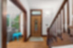 Bright home entryway with wooden staircase, front door, and cozy nook with gray chair and blue table by the window.