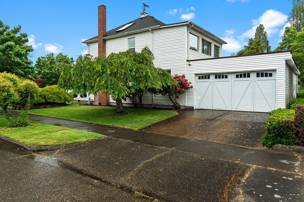 White two-story house with two car garage and brick chimney on a wet driveway, surrounded by lush green trees under a blue sky