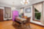 Bright dining room with wooden table and chairs, pink cabinet, beige curtains, floral chandelier, and framed landscape art.