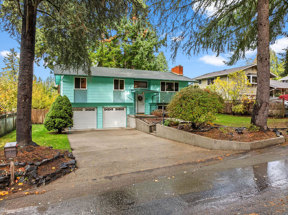Turquoise house with two garage doors and a driveway, surrounded by trees and shrubs. Blue sky and neighboring house in the background.