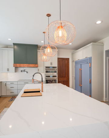 View of quartz island with sink, range and hood, pendant lighting and refrigerator built-ins in kitchen with copper accents