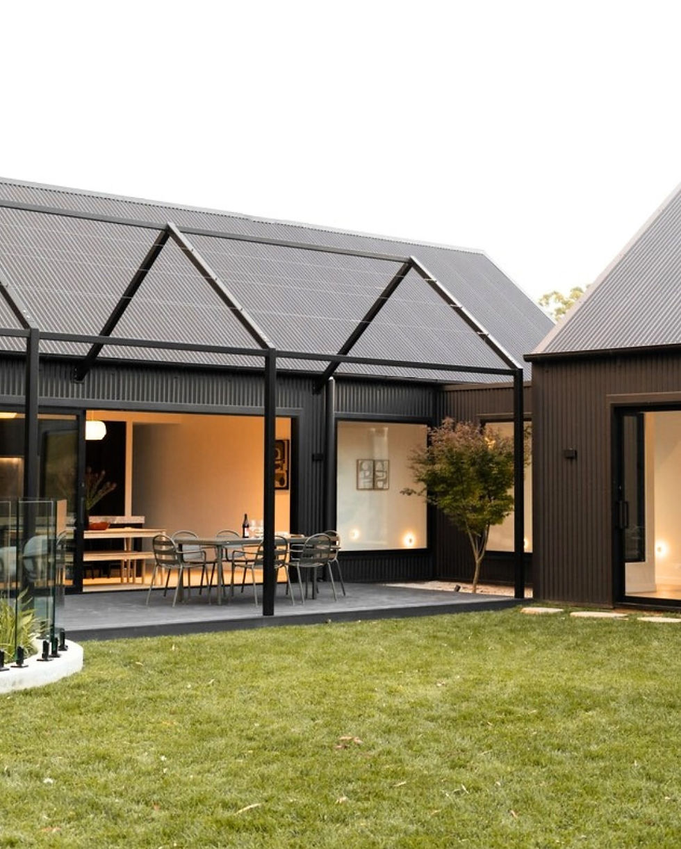 Modern house exterior with dark cladding, glass doors, outdoor dining set on a patio, and a manicured lawn, creating a cozy evening ambiance.