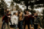 Group of smiling people in a forest, giving piggyback rides. A white van and wrapped gifts are in the background, creating a festive mood.