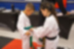A martial arts instructor ties a green belt on a child in a dojo. Both wear white uniforms. Red and black mats cover the floor.
