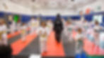 Children in white martial arts uniforms with colored belts practice in a gym. Instructor in black supervises. Bright, organized setting.