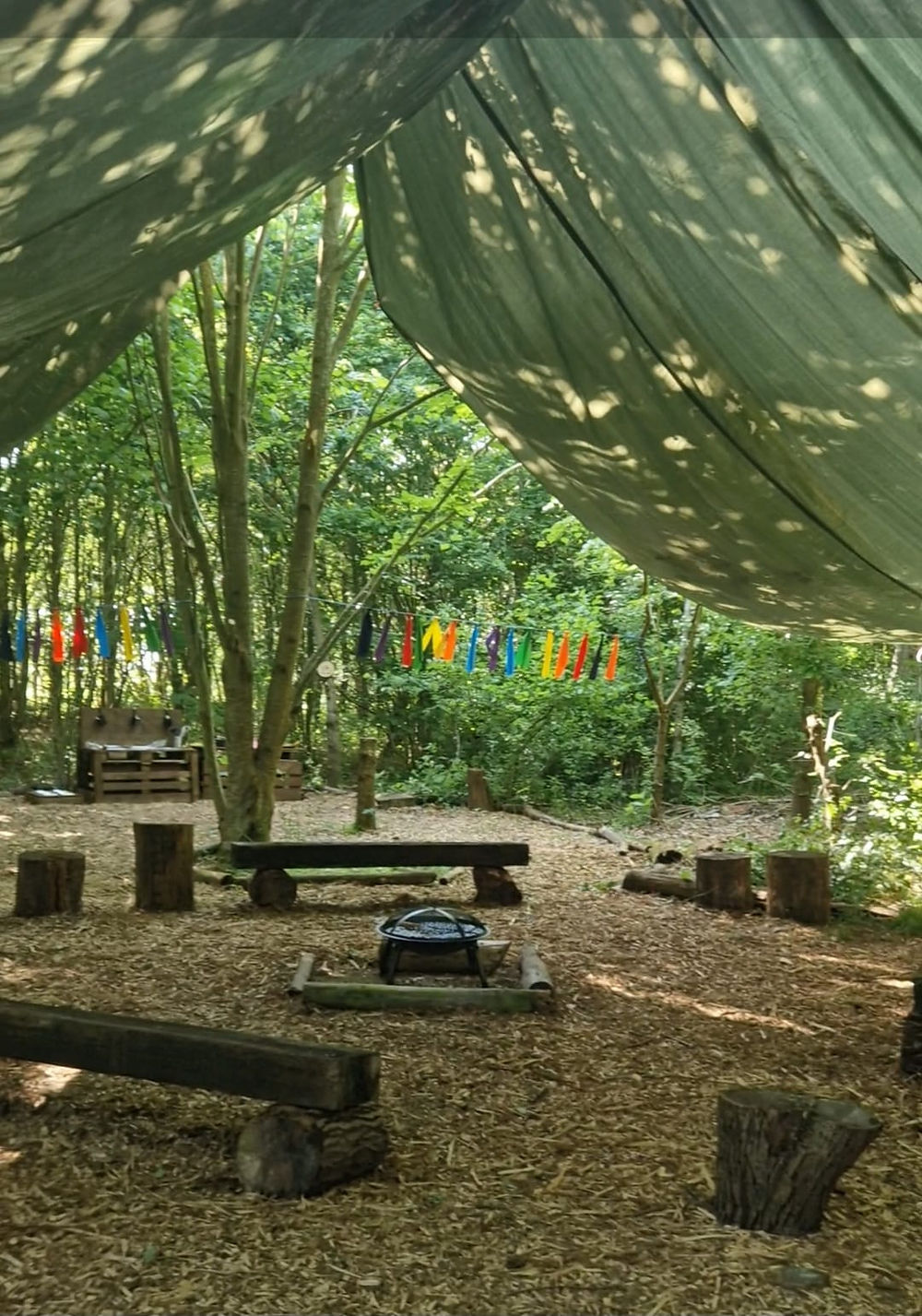Wildbound forest school basecamp. A campfire beneath a large tarp surrounded by logs