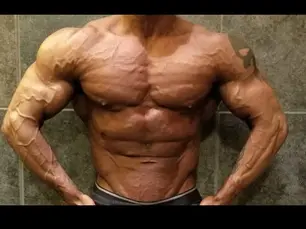 Bodybuilder flexing muscles in a gym, showing a muscular, defined torso. Gray tiled background, arms positioned on hips, intense focus.