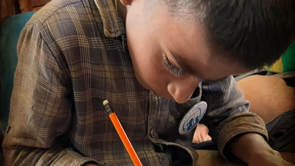 Young boy in a plaid shirt focuses on drawing with an orange pencil. He's seated indoors, with concentration visible on his face, creating a quiet and studious mood.