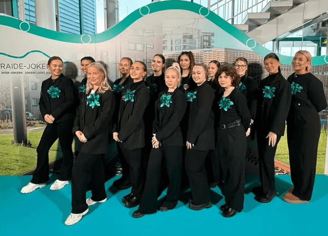 Ryhmää mustissa asuissa, joissa turkoosit kukkakoristeet, poseeraa sisätiloissa kirkkaansinistä mattoa vasten. Taustalla mainosjuliste.