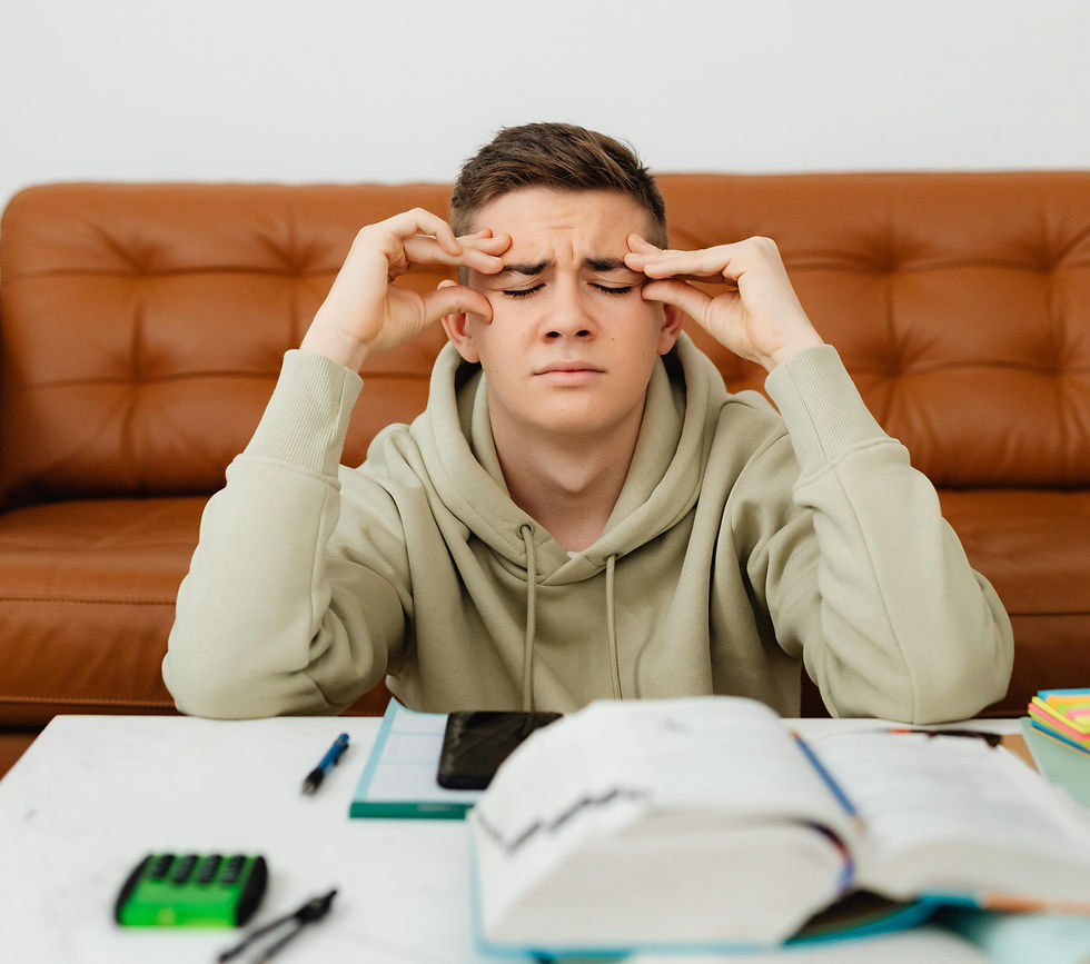Teen in a light hoodie sits at a table with open books, pens and a calculator, rubbing temples, appearing stressed. Brown sofa in background.