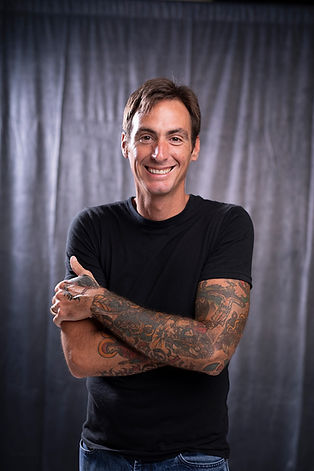Michael Beck with a gray background. He is smiling, crossing his tattooed arms, and wearing a black tshirt and jeans.