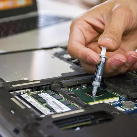 A close-up shot of a technician’s hand using a syringe to apply a precise amount of grey thermal compound onto a central processing unit during a laptop motherboard repair.