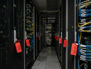 Narrow data center aisle lined with black server racks and blue/yellow cables, red warning tags hanging on doors, dimly lit.