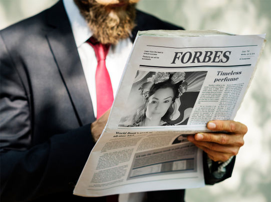 A man in a suit holds a Forbes newspaper. On the right page of the newspaper, there is a photo of Olga-Alaia Mariná and an article about her successful business — the niche perfume 'ALAIA perfume.'