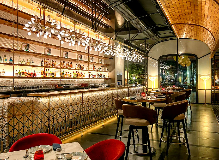 The heart of KOKO is its grand bar setup, and the rest of the venue naturally flows around it. Spanning three floors, the space shifts from an airy, plant-filled outdoor patio on the ground level to a high-energy lounge above. The structure features an exclusive, private sixteen-seat room tailored for intimate gatherings, while the top floor opens up into a sweeping terrace under the stars. Linear design details, soft lighting, and plush seating work together to keep the vast layout feeling warm and connected. Downstairs, the mood softens with lower lamps that make tables feel private, though the central seating stays lively and close to the action. It is among the best bars in Bengaluru.