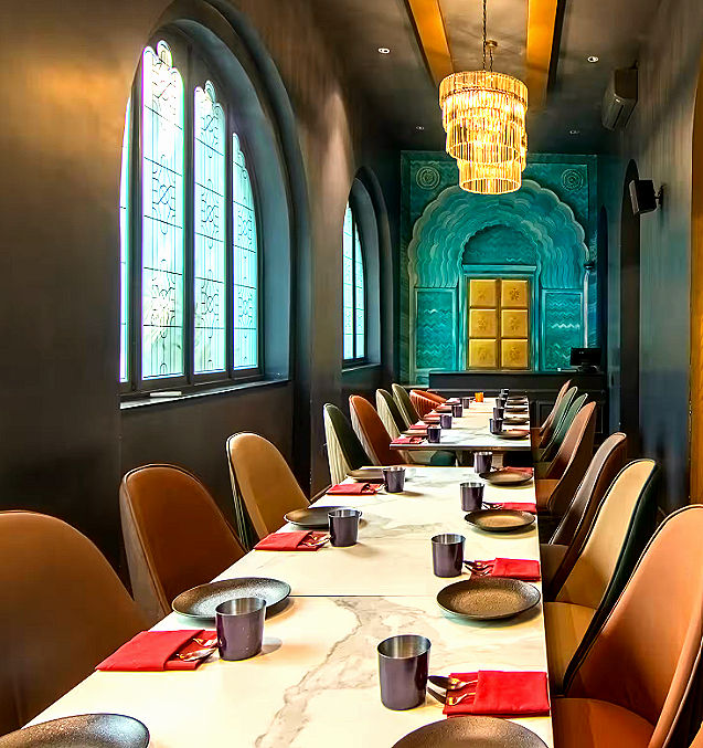 The interior features monochrome geometric marble tile floors, dramatic deep-crimson wall panelling, and plush velvet armchairs reminiscent of a modern royal parlour. Custom brass orb chandeliers suspended overhead cast a warm, focused glow over every place setting, transforming each course into a visual showcase. Elegant archways, hand-carved accents, and bespoke art murals frame the dining hall, offering a rich, sophisticated backdrop that enhances the vivid colors of progressive Indian plating.