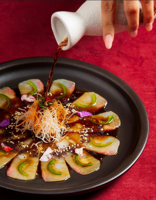 The food program at KOKO elevates classic Cantonese and Japanese recipes by pairing precise traditional techniques with luxurious raw ingredients. The cold kitchen centers on delicate seafood preparations, showcasing their Hamachi Carpaccio sliced thin and dressed in a bright, citrusy yuzu-soy base with jalapeño. The sushi selection features complex rolls like the Chili Salmon Roll and the black rice edamame roll, highlighting varied textures and clean sea-salted flavors. Hot dishes transition into rich, steamed dim sum such as the lobster and caviar dumpling. Delicate starches are utilized to produce glass-like dumpling wrappers and thin sheets of cheung fun, ensuring the natural sweetness of the seafood remains the dominant note here among the Old Airport Road bars.