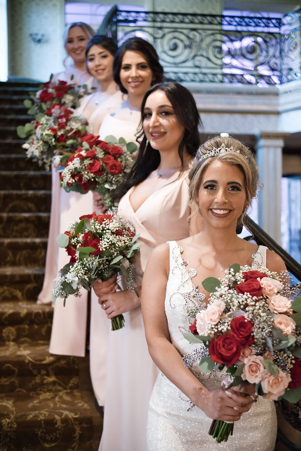 Wedding Photographer in NJ | Bridesmaids in pastel dresses hold red and pink bouquets, standing on ornate stairs with smiles. Elegant venue, joyful mood.