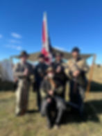 Five Civil War reenactors pose with rifles before a Confederate flag and tents under a bright blue sky.