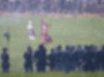 Civil War reenactors in blue uniforms stand with rifles and flags in a smoky grassy field, with Confederate reenactors behind a dirt berm.