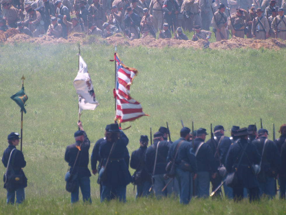 Civil War reenactors in blue uniforms stand with rifles and flags in a smoky grassy field, with Confederate reenactors behind a dirt berm.