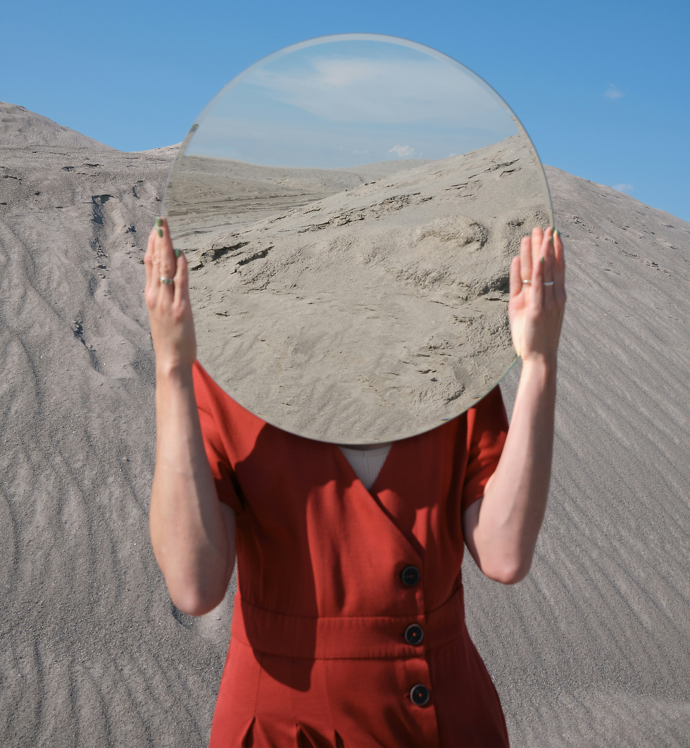 Woman in a red dress standing on sand dunes holding a round mirror in front of her face reflecting the sky.