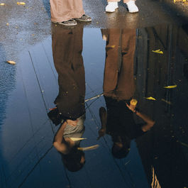 Two people standing on a wet street next to a puddle, with their full reflections visible in the water.