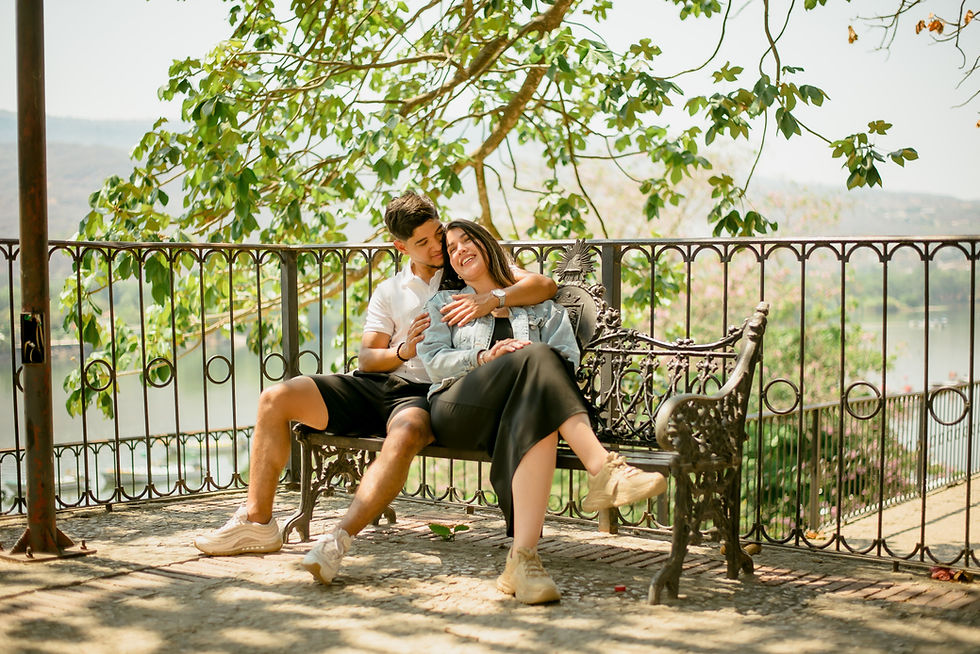 Smiling couple cuddles on an ornate iron bench under leafy trees, overlooking water and hills in a sunny park.