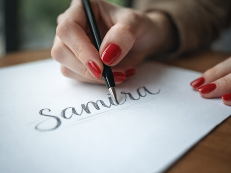 Close-up of a woman's hand with long red polished nails sketching the name 'Samira' in cursive on white paper, symbolizing personal logo design and creativity.