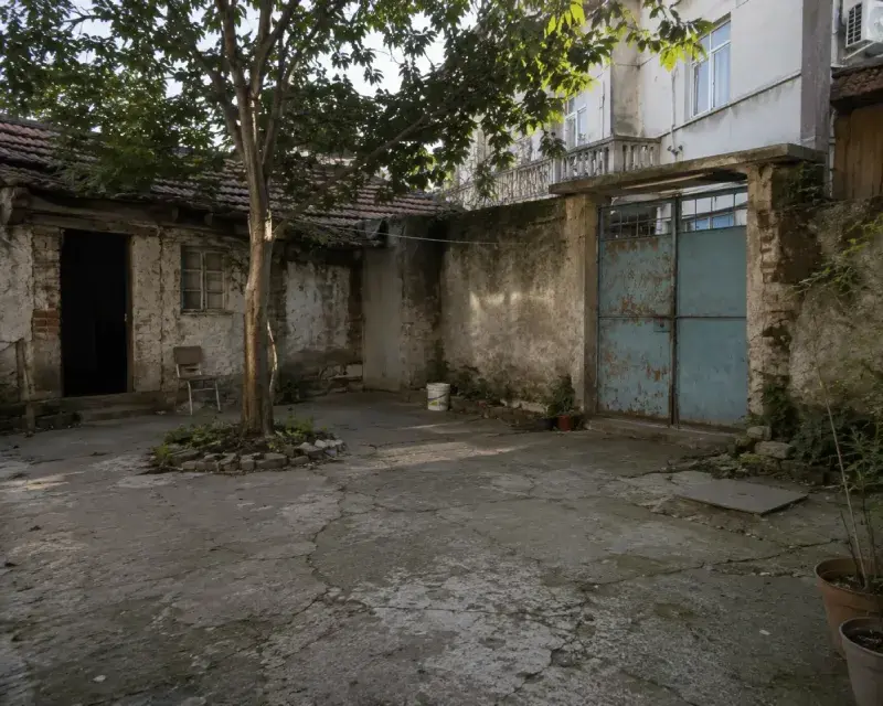 Photo of an old, weathered courtyard with cracked concrete paving, aged masonry walls, a rusted blue metal gate, a mature tree in the center, and a neglected outdoor space awaiting landscape renovation.