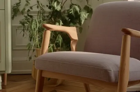 Close-up of a wooden armchair with light fabric upholstery in a softly lit interior with plants.