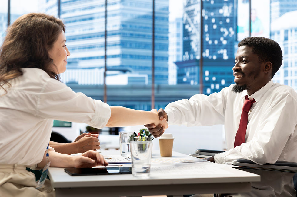 Two people in business attire, smiling and shaking hands across a desk in a modern office with large windows and city view.