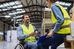 Man in wheelchair with tablet, smiling at a female colleague with clipboard. Both in yellow vests, inside a bright warehouse.
