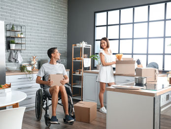 Smiling couple unpacking boxes in a bright modern kitchen, with a man in a wheelchair holding a box.