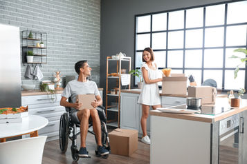 Smiling couple unpacking boxes in a bright modern kitchen, with a man in a wheelchair holding a box.