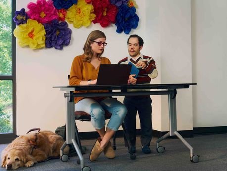 Woman sits at a desk as a man stands next to her. The two look at a laptop. A service dog sits at the woman's feet.