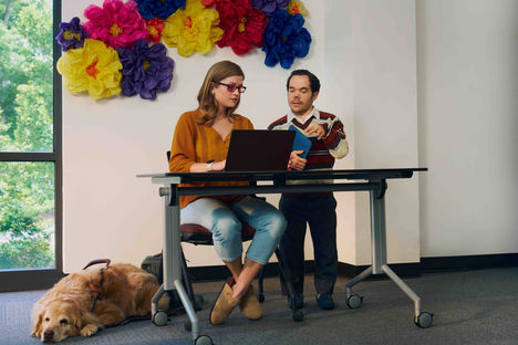 Woman sits at a desk as a man stands next to her. The two look at a laptop. A service dog sits at the woman's feet.