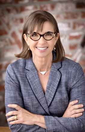 Woman with glasses and a gray blazer smiles, arms crossed. Brick wall background. Professional and confident mood.