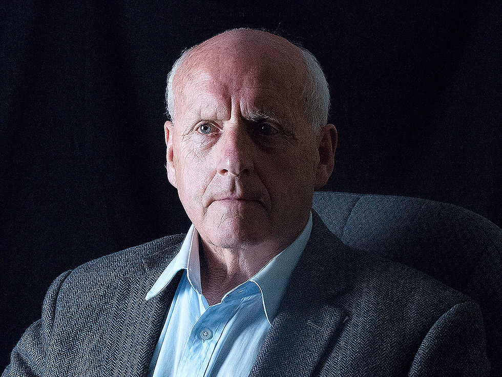Serious bald older man in a gray blazer and white shirt, seated against a dark studio background, lit from the left.