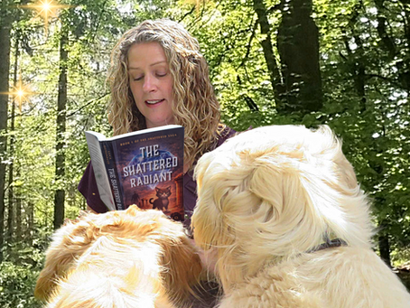Woman reading The Shattered Radiant in a sunlit forest while two fluffy dogs nuzzle her.