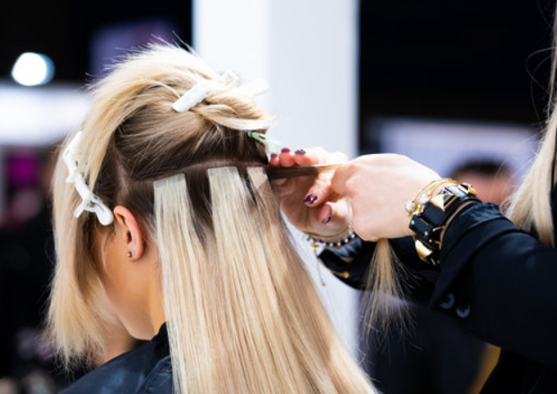 Hair stylist adds blonde Tape in Hair extensions to a woman’s hair in a salon, with clips and tools visible.