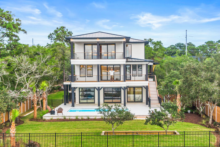 Wide rear view of elevated coastal custom home with pool patio, porches, and fenced backyard.