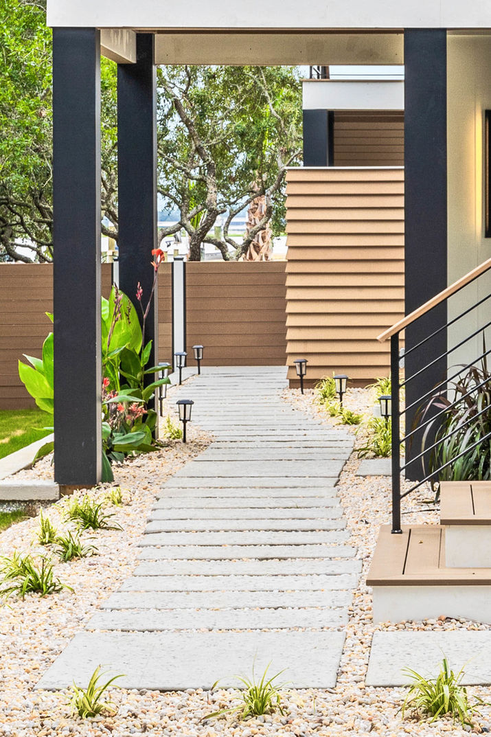 Modern coastal walkway with concrete pavers, gravel landscaping, black columns, and exterior lighting.