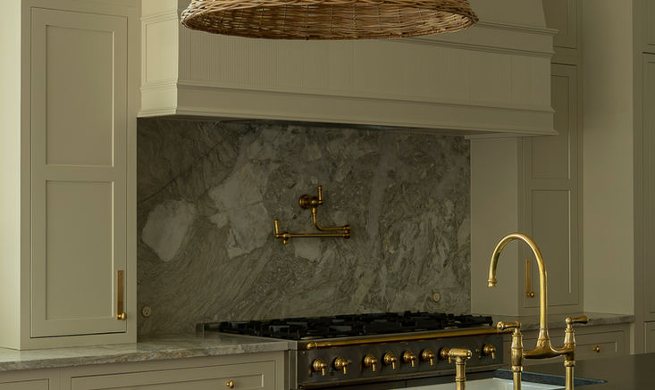 Custom kitchen design featuring a warm wood island with black countertop, soft neutral cabinetry, marble backsplash, brass fixtures, and oversized woven pendant lighting.
