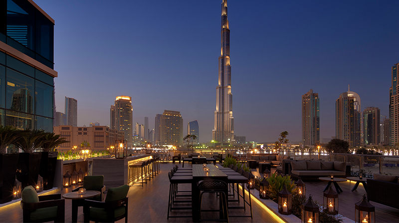 Rooftop dining area with tables, lanterns, and greenery overlooking a city skyline at dusk. Notable skyscraper in the background. Calm ambiance.
