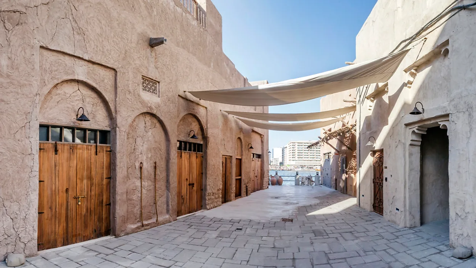 A rustic alley with arched doorways and wooden doors, shaded by cloth canopies. Sunlit, with a modern city view in the distance.