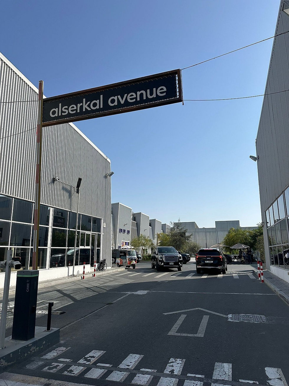 Street view of Alserkal Avenue sign above, with cars and a tuk-tuk on the road. Industrial buildings and a clear blue sky in the background.