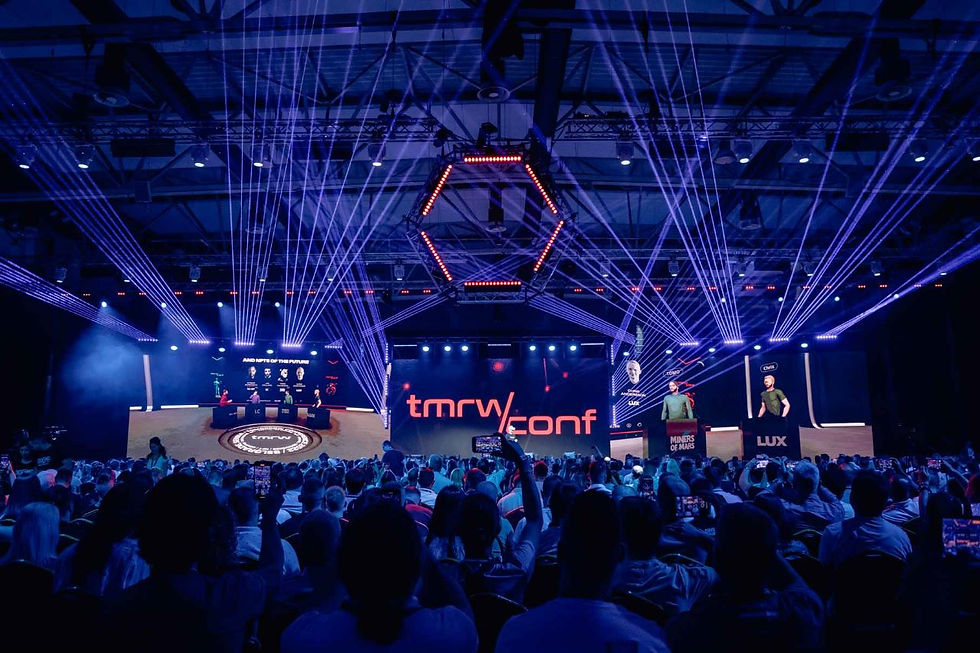 Audience watches a vibrant stage with purple lights at tmrw conf. Speakers on stage under geometric lighting. Mood is energetic and tech-focused.