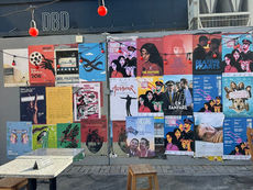 Street scene with colorful posters on a wall, red string lights above. Three wooden stools and a small table below, creating an urban vibe.