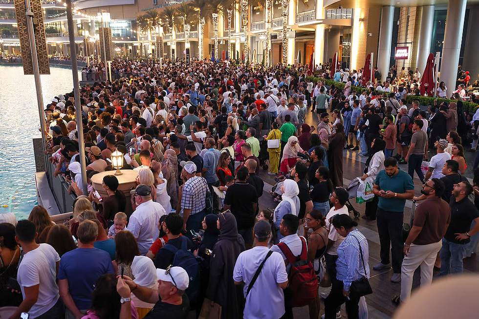Crowd of people gathered by a waterfront at sunset. Bright lights illuminate a building with palm trees in the background, creating a lively atmosphere.